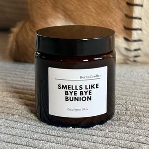 Könnte beinhalten: Ein dunkelbraunes Glas mit schwarzem Deckel. Das Etikett lautet "Smells Like Bye Bye Bunion" und "Eucalyptus Glow". Das Glas steht auf einer grauen Oberfläche, mit unscharfem Hintergrund.