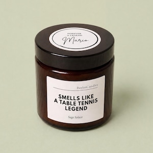 Peut inclure: Un bougeoir en verre marron avec un couvercle noir. L'étiquette indique "Smells Like A Table Tennis Legend" et "Sage Solace". Le couvercle porte l'inscription "Forever A Legend" et le nom "Mario".