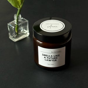 Puede incluir: Un tarro de vela de vidrio marrón con una tapa negra y una etiqueta blanca que dice "Smells Like A Future Lawyer". Un pequeño jarrón de vidrio con una planta verde está a la izquierda de la vela.