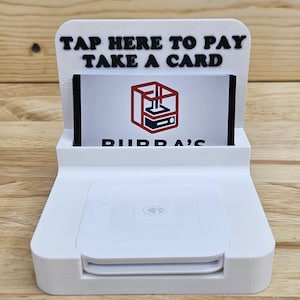 May include: A white contactless payment stand with the text "TAP HERE TO PAY TAKE A CARD" in black. The stand holds a card with a logo and the text "PIIDDA'S". The stand has a wireless symbol.