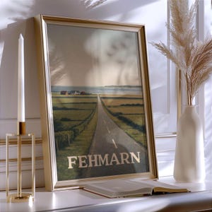May include: A framed print featuring a long road leading to the sea, with the word "FEHMARN" in gold lettering. The artwork is in a gold frame, and is propped up on a white surface. A white candle and a vase with dried plants are also visible.