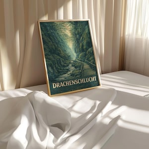 May include: A framed print featuring a scenic landscape of a gorge with a wooden bridge and a river. The artwork is dominated by shades of green and brown, with the text "DRACHENSCHLUCHT" at the bottom. The frame is gold.