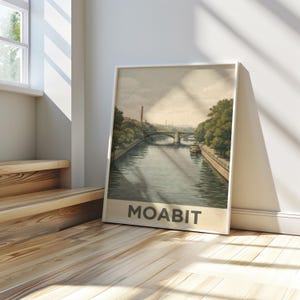 Puede incluir: Una impresión artística enmarcada que presenta una ilustración vintage de una escena fluvial con un puente, árboles y un barco. La impresión tiene un borde blanco y la palabra "MOABIT" en la parte inferior. La obra de arte se exhibe en un suelo de madera.