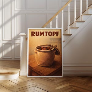 May include: A framed print with the word "RUMTOPF" at the top. The image shows a ceramic pot with a lid, filled with dark fruit. The pot is on a wooden table, set against a warm, soft background.