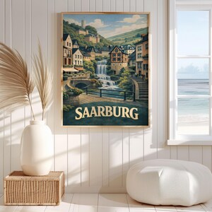 May include: A framed wall art print depicting a scenic landscape of Saarburg, Germany, featuring a multi-tiered waterfall flowing through a town with half-timbered buildings, a stone bridge, and the text SAARBURG in bold, gold-coloured lettering.