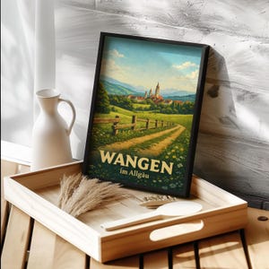 Könnte beinhalten: Gerahmter Kunstdruck mit einer Landschaftsdarstellung mit einem Weg, der zu einem Dorf führt. Der Druck hat einen schwarzen Rahmen und den Text "WANGEN im Allgäu" in Gold. Er wird auf einem Holztablett mit dekorativen Elementen präsentiert.