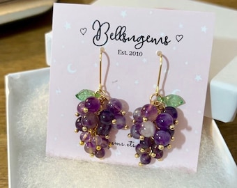 May include: A pair of amethyst grape earrings with gold wire and green leaf accents. The earrings are displayed on a pink card with the text "Bellisgems Est. 2010".