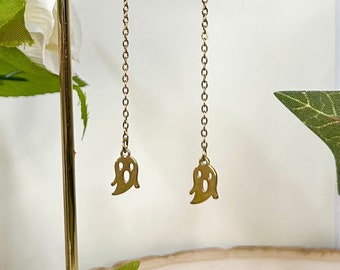 May include: Gold-colored dangle earrings featuring a chain design with a small ghost charm at the bottom. The earrings are displayed on a gold-colored earring stand against a white background with greenery.