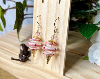 May include: A pair of dangle earrings with a whimsical ice cream cone design. The earrings feature pink and white striped glass beads with red polka dots, gold-colored cone bottoms, and gold-tone ear wires. Clear and pink crystal beads are at the top.