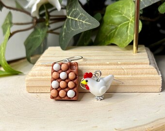 May include: A pair of earrings featuring a carton of eggs and a white chicken. The egg carton is brown with white and brown eggs. The chicken has a red comb and yellow beak. The earrings are displayed on a wooden stand.