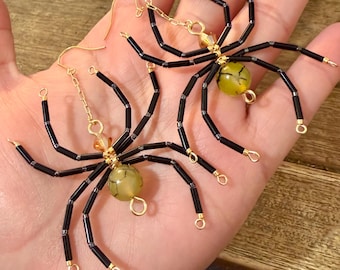 May include: A pair of spider-shaped earrings featuring black tubular glass beads for legs, a central gold-toned wire frame, and a round, crackled light green stone bead for the body, suspended from gold-toned metal earring hooks.