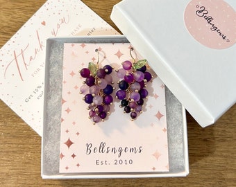 May include: Purple and green grape cluster earrings with gold accents. The earrings are displayed in a white box with a pink and white card that says "Bellsngems Est. 2010".