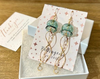 May include: A pair of green glass and gold dangle earrings with a white pearl accent. The earrings are displayed on a white card with the text "Bellsingers Est 2016".