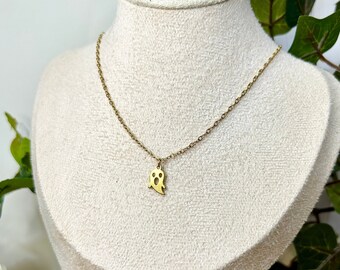May include: A gold-colored necklace with a small ghost pendant. The pendant is a simple cutout design. The necklace is displayed on a mannequin bust, with green foliage in the background.