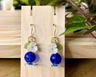 May include: A pair of handmade earrings featuring a gold-colored hook, a blue bead, a white flower, and green leaves. The earrings are displayed on a wooden stand against a backdrop of greenery.