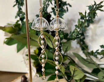 May include: A pair of gold dangle earrings with clear glass bell-shaped charms and white pearl beads. The earrings are hanging from a gold stand.