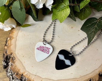 May include: Two guitar picks on silver chains. One pick is white with pink text that says "Arctic Monkeys". The other pick is black with a white sound wave graphic.