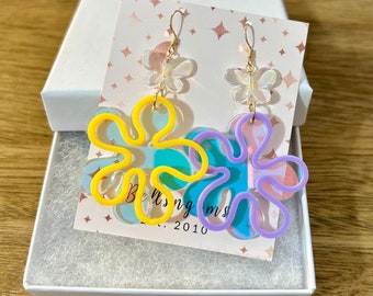 May include: A pair of colorful flower earrings with gold-colored hooks. One earring is yellow, and the other is purple. Each earring has a clear butterfly charm. The earrings are displayed in a white gift box.