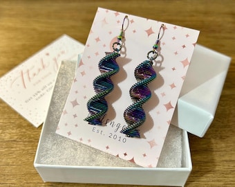 May include: A pair of iridescent spiral earrings with a dark metal finish, displayed in a white gift box. The earrings have a unique, twisted design and are presented on a pink card with the text "lsnge Est. 2010".