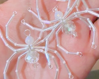 May include: Three spider-shaped ornaments crafted from iridescent white tube beads and clear faceted beads. Each spider features a central body made of a round bead and eight spindly legs constructed from segments of white, shimmering glass-like beads.