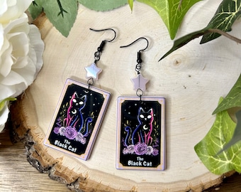 May include: A pair of black cat tarot card earrings with a silver star charm. The cards are black with a pink and blue cat illustration and the text "The Black Cat".