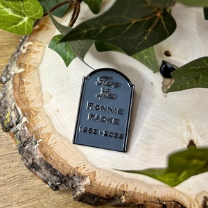 May include: Black enamel pin shaped like a tombstone with the text "Here Lies Ronnie Radke 1983-2023".