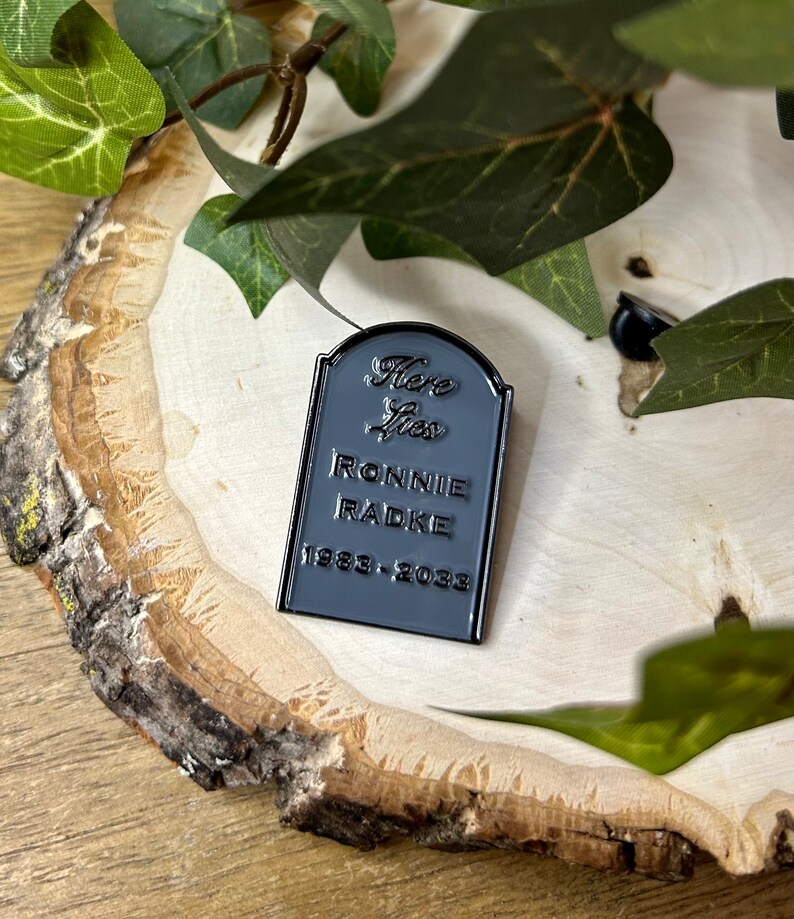 May include: Black enamel pin shaped like a tombstone with the text "Here Lies Ronnie Radke 1983-2023".