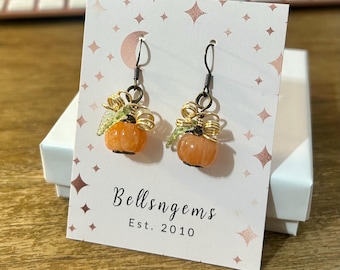 May include: A pair of dangle earrings featuring small orange pumpkin charms with gold-toned wire coils and green leaf accents. The earrings hang from dark metal hooks on a white display card printed with the text Bellsngems Est. 2010.
