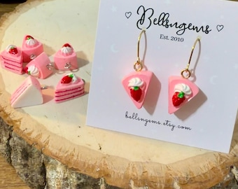 May include: A pair of dangle earrings shaped like pink strawberry cake slices with white frosting and a red strawberry on top, attached to a white display card with the text Bellsugems Est. 2010 and bellsngems.etsy.com, alongside loose cake charms.