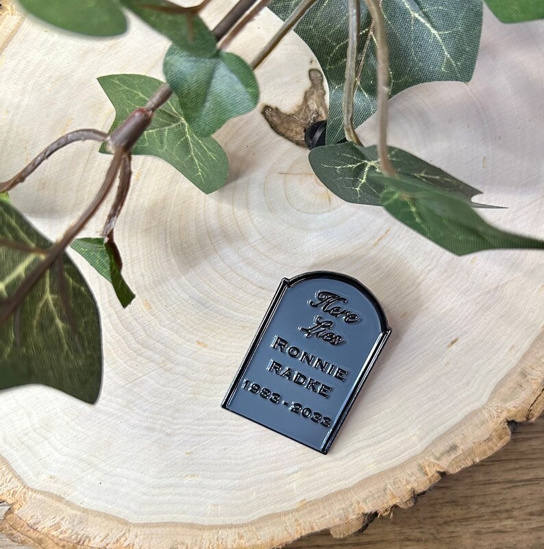 May include: Black enamel pin shaped like a tombstone with the text "Here Lies Ronnie Radke 1982-2023".