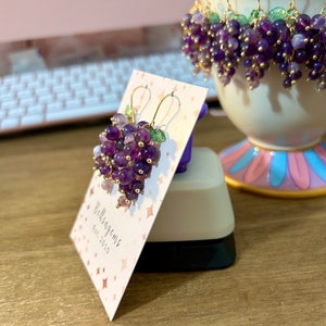 May include: A pair of amethyst grape earrings with gold wire and green leaves. The earrings are displayed on a white card with the text "Bellisagems Est. 2010".