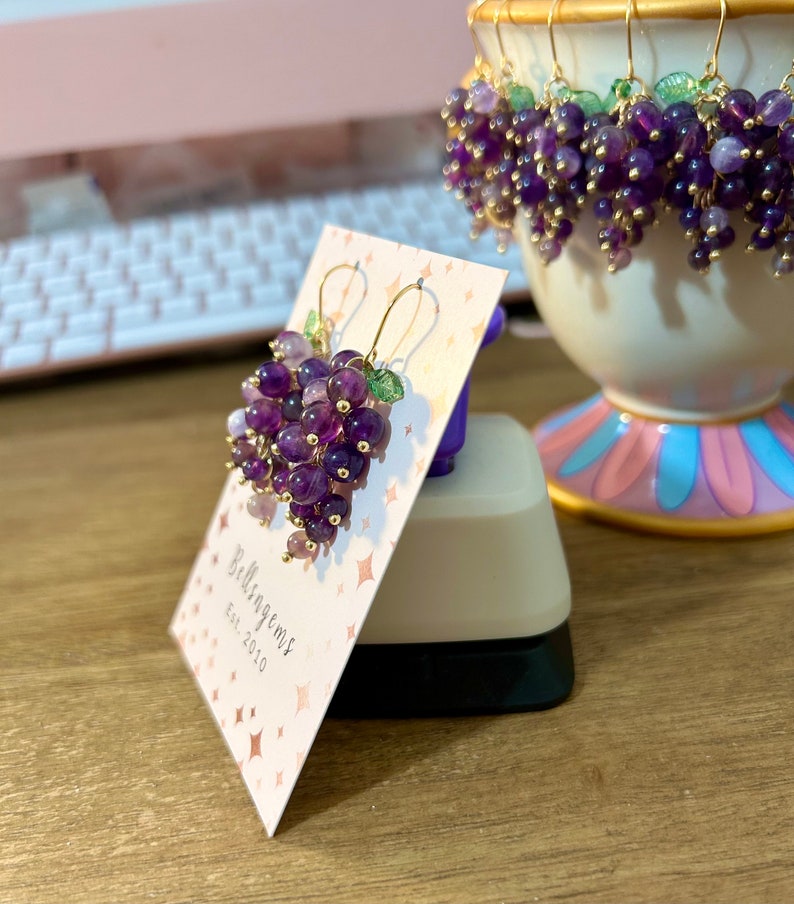 May include: A pair of amethyst grape earrings with gold wire and green leaves. The earrings are displayed on a white card with the text "Bellisagems Est. 2010".