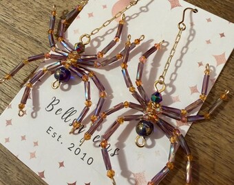 May include: A pair of spider-shaped earrings crafted from purple and orange beads, featuring gold-toned metal chains and hooks. The earrings are displayed on a white card printed with the text 'Bells Gems Est. 2010' and a pattern of small stars.