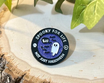 May include: A round, black enamel pin with the text "Spooky for Life, Not Just Halloween." The pin features a white ghost, bats, and the word "Boo" in a speech bubble against a purple starry background. Two white skulls are on either side.