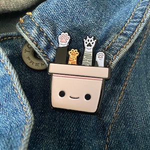 May include: A whimsical enamel pin featuring a smiling planter pot with four cat paws in various colors. The paws are pink, orange, and white, set against a black and white pot. The pin is attached to a denim jacket.