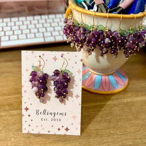 May include: A pair of amethyst grape earrings with gold-toned hooks and green leaves. The earrings are displayed on a card with the text "Bellsngems Est. 2010".