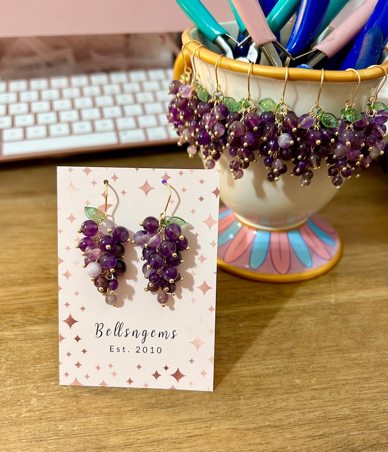 May include: A pair of amethyst grape earrings with gold-toned hooks and green leaves. The earrings are displayed on a card with the text "Bellsngems Est. 2010".