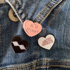May include: Three enamel pins on a denim jacket. The pins are heart-shaped. One pin is pink and says "RU Mine?" Another pin is black and has a waveform design. The third pin is white and says "Arctic Monkeys".