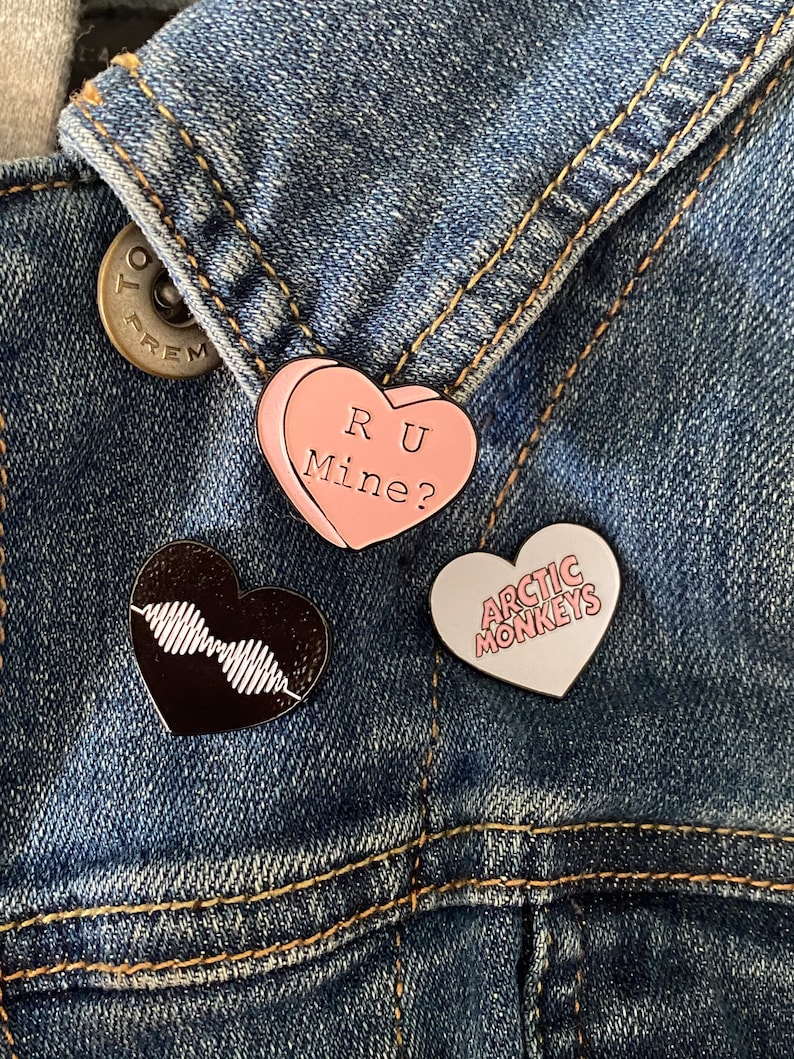 Puede incluir: Tres pines de esmalte en una chaqueta vaquera. Los pines tienen forma de coraz&oacute;n. Un pin es rosa y dice "RU Mine?" Otro pin es negro y tiene un dise&ntilde;o de forma de onda. El tercer pin es blanco y dice "Arctic Monkeys".