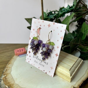 May include: Amethyst grape earrings with gold wire and green leaves. The earrings are displayed on a pink card with the text "Bellsngems Est. 2010".
