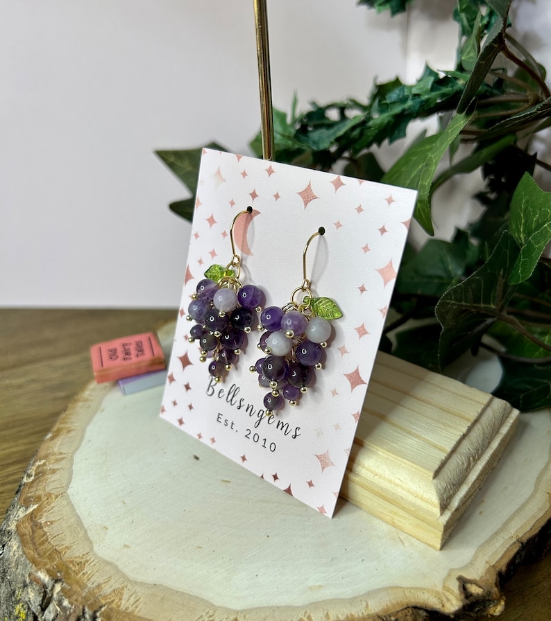 May include: Amethyst grape earrings with gold wire and green leaves. The earrings are displayed on a pink card with the text "Bellsngems Est. 2010".