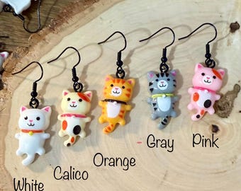 May include: Five cat-shaped dangle earrings with black metal hooks. The cats are white, calico, orange, gray, and pink, each featuring a small collar. Text labels below each earring identify the colors as White, Calico, Orange, Gray, and Pink.