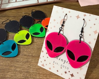 May include: A collection of alien-shaped dangle earrings in neon pink, neon green, bright blue, and black. Each earring features a cutout design with black eye details and black metal hooks. The pink pair is displayed on a white card with star patterns.