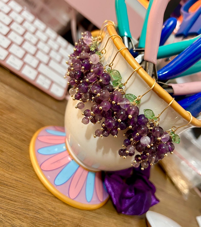 May include: Three pairs of dangle earrings with amethyst and green glass beads, shaped like bunches of grapes, hanging from gold-toned hooks.
