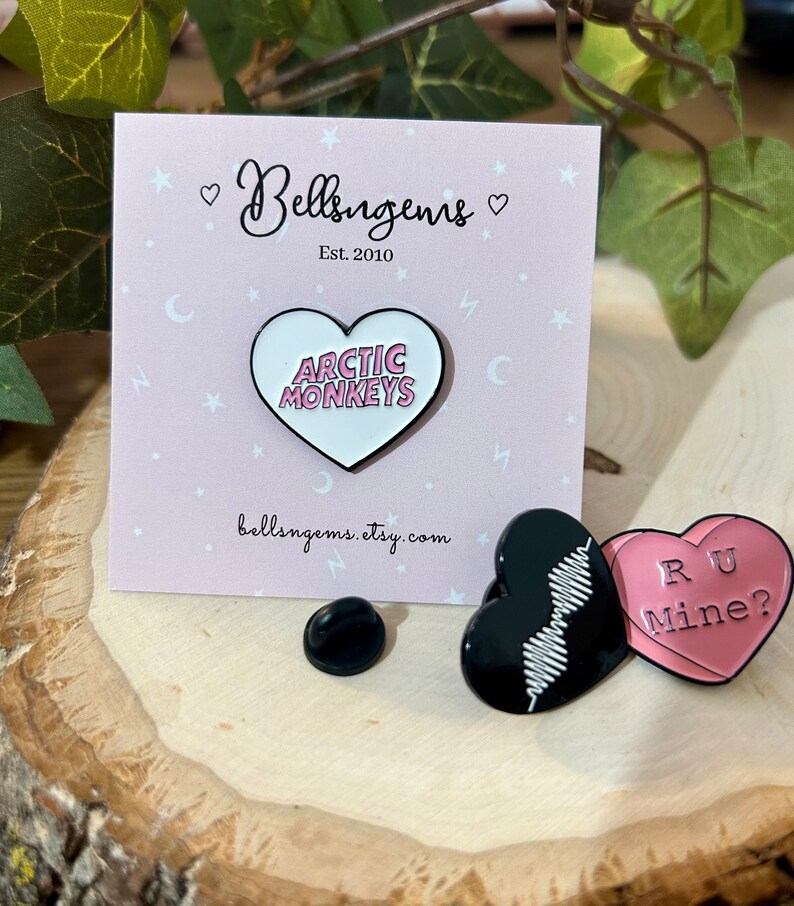 Puede incluir: Dos pines de esmalte con la banda Arctic Monkeys. El primer pin es un coraz&oacute;n rosa con el nombre de la banda en letras blancas. El segundo pin es un coraz&oacute;n negro con un patr&oacute;n de onda blanca y las palabras "RU Mine?" en letras rosas.