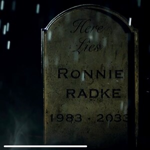 May include: A weathered grey tombstone with the inscription "Here Lies Ronnie Radke 1983 - 2033" in black lettering. The tombstone is set against a dark background with rain falling.