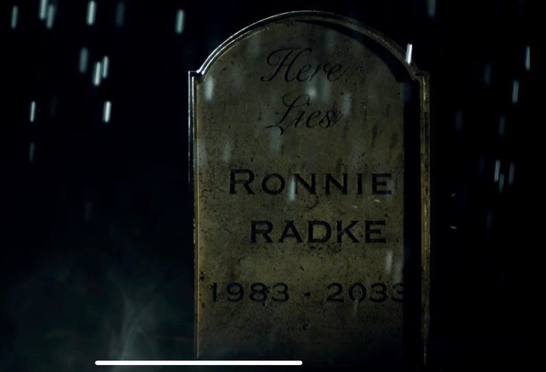 May include: A weathered grey tombstone with the inscription "Here Lies Ronnie Radke 1983 - 2033" in black lettering. The tombstone is set against a dark background with rain falling.