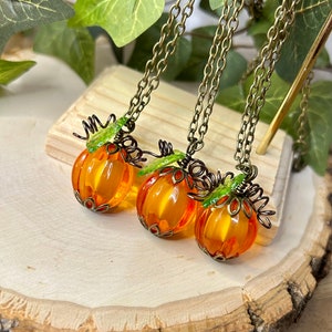 May include: Three pumpkin-shaped necklaces with orange glass beads and bronze-colored metal accents. Each necklace has a bronze chain and green leaf details. The necklaces are displayed on a wooden surface, with green leaves in the background.