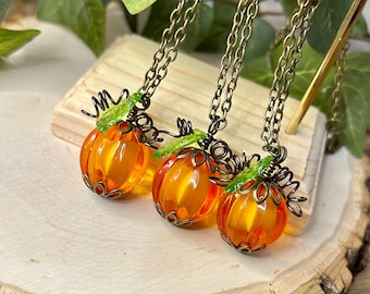 May include: Three pumpkin-shaped necklaces with orange glass beads and bronze-colored metal accents. Each necklace has a bronze chain and green leaf details. The necklaces are displayed on a wooden surface, with green leaves in the background.
