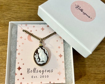 May include: A white cameo necklace with a gold chain and a brown oval pendant featuring a white cat and a rabbit. The pendant is in a white box with a pink and white star pattern and the text "Bellsggems Est. 2010".